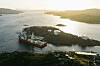 Luftfoto av stort rødt konstruksjonsskip ved kai på en øy omgitt av fjord og lagertanker.