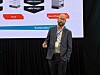 En mann står på en scene foran en skjerm med illustrasjoner av batterimoduler og tekst om energiløsninger.