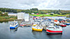 Ulike båter ligger fortøyd ved et verft med industribygg og en rolig fjord i forgrunnen.