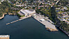 Flyfoto av et havneområde med kaier, industribygg, sjø og boligområder i kupert terreng.