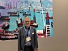 En mann med navneskilt står foran en illustrasjon av skip, havn og kraner på en maritim messe.