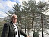 Mann i dress står foran furutrær ved en fjord under en delvis skyet himmel.