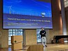 Person presenterer foran en stor skjerm med tekst og illustrasjon av havvindmøller i en konferansesal.